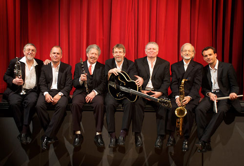 7 Mitglieder der Barrelhouse Jazzband sitzen auf der Bühne, im Hintergrund ein roter Vorhang.
