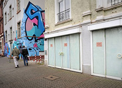Zwei Fußgänger auf der leeren Berger Straße. Im Vordergrund verklebte Fensterscheiben eines geschlossenen Geschäfts.